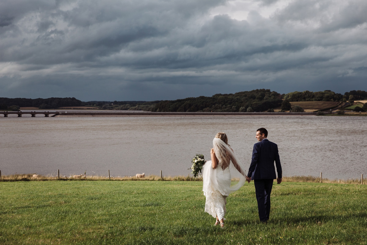 Bride and Groom stroll by the waters edge with a stormy sky overhead at Blithfield Lakeside Barns